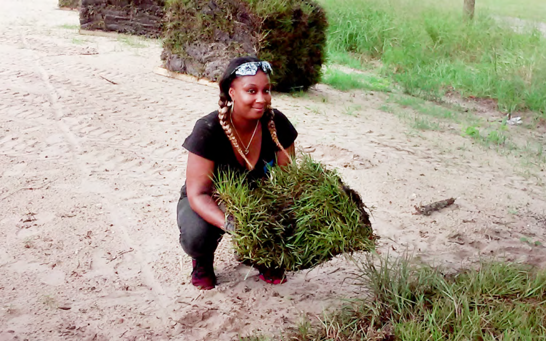 Woman smiling crouched down holding sod on sand lot