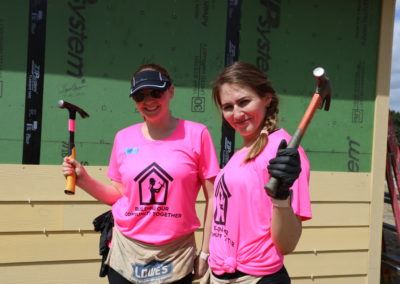 Two women wearing pink shirts and holding hammers in front of in-progress siding of house