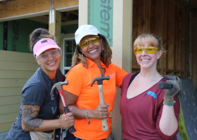 Three smiling women holding hammers and wearing safety goggles in front of in-progress house