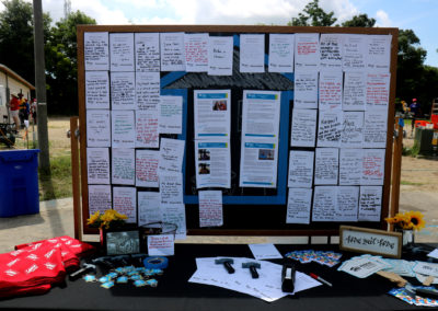 Bulletin board with handwritten papers and blue taped outline of a house behind a table with foam hammers, papers, and key chains.