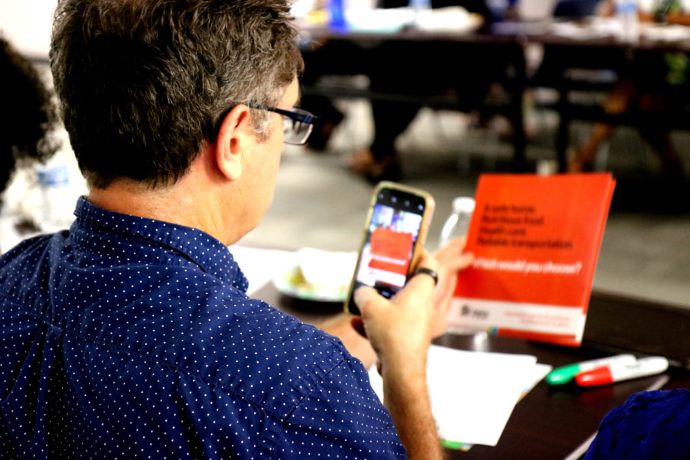 Man wearing polka dot shirt taking a photo on his phone of orange paper with wording on it