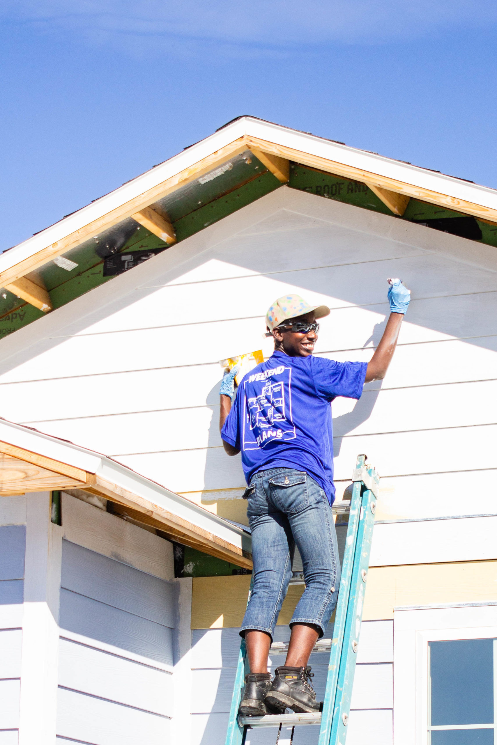 Smiling volunteer standing on a ladder and painting siding on front of house